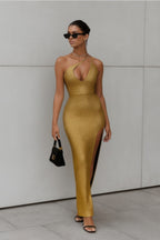 Woman wearing a gold dress with a high slit, standing against a gray wall.